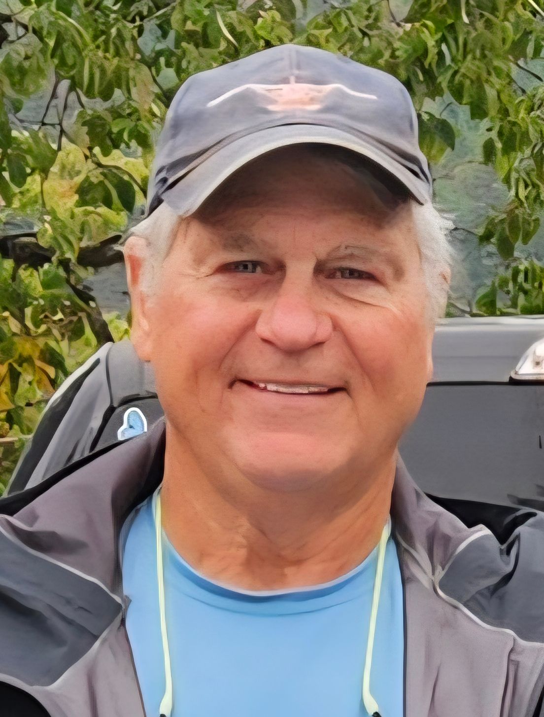 Man wearing a baseball cap smiles, outdoors with trees in the background.