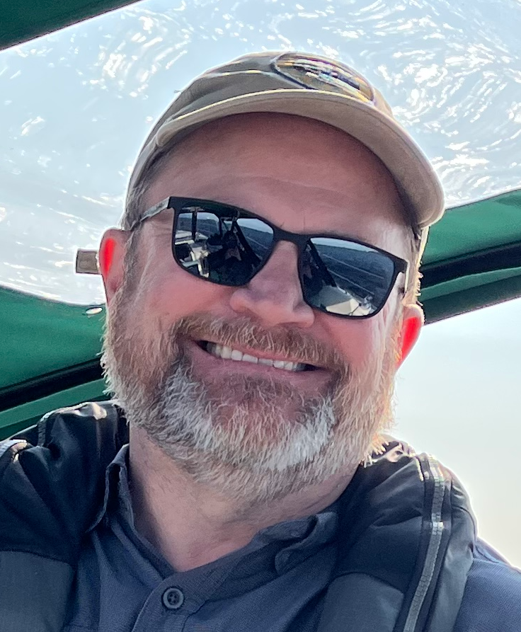 Man wearing sunglasses and a cap, smiling on a boat.