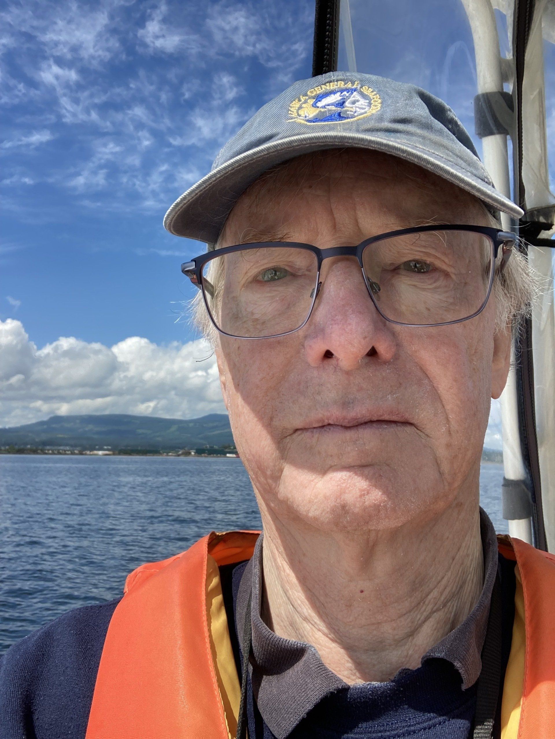 Man in a hat and glasses on a boat, wearing a life vest. Ocean and land in the background.
