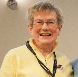 Woman with short hair, wearing glasses and a yellow top, smiling. Black lanyard around neck.