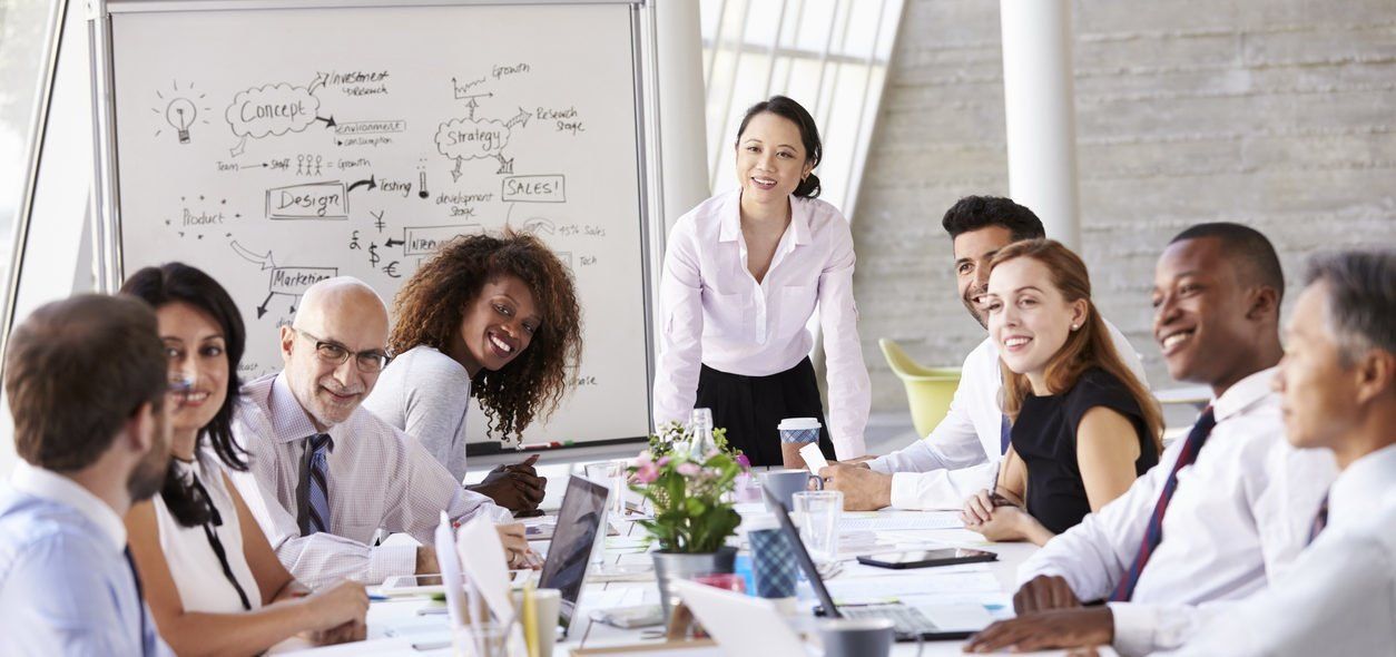 Equipe de 8 executivos sentados ao redor de uma mesa de trabalho e uma jovem mulher em pé na lateral da mesa em frente a um quadro branco