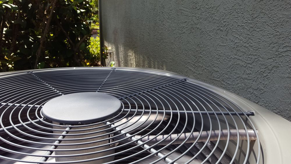Close-up of an outdoor air conditioner unit with a metal grill fan. Grey stucco wall and foliage in the background.