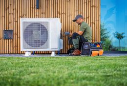 A person in work clothes repairs an AC unit outside a modern building.