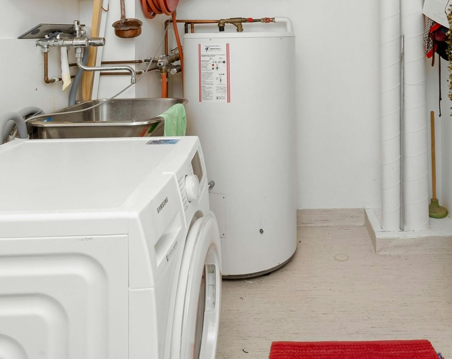 Laundry room with washing machine, water heater, and sink. White appliances and walls, small red rug.