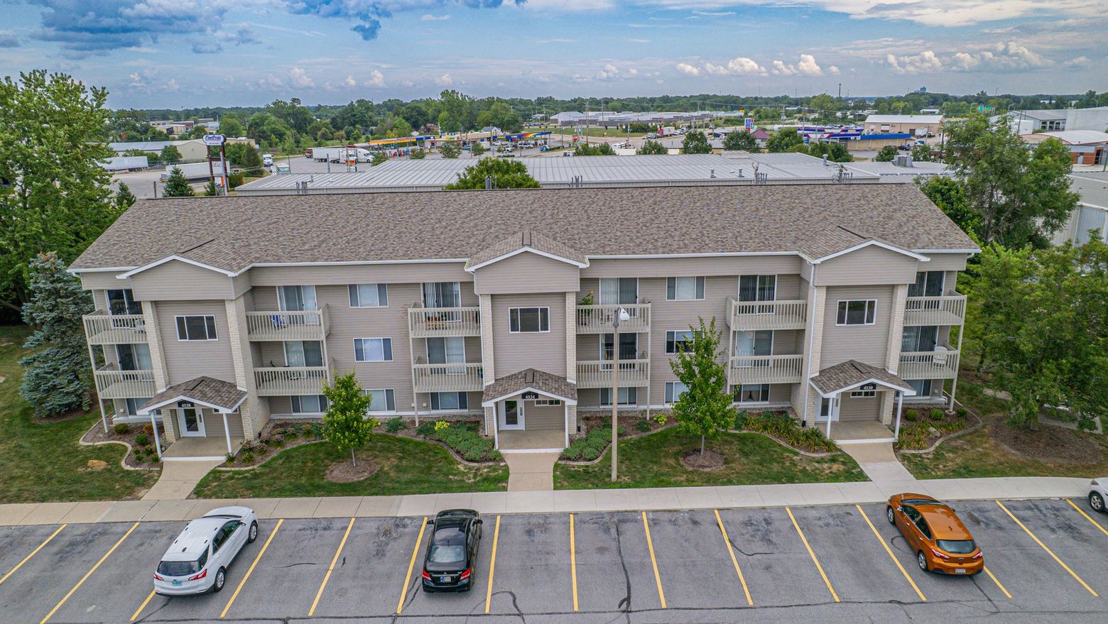 Aerial view of a beige apartment building with balconies, parking lot in front, and surrounding trees.