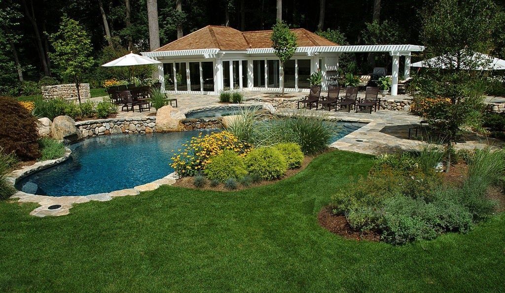 Swimming pool and patio area with a house in the background; lush green lawn, blue water.