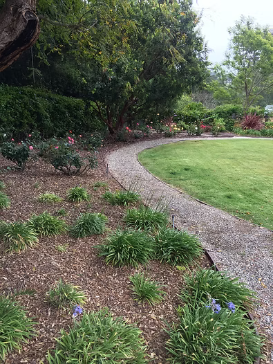 Gravel path curves through a garden with green grass, shrubs, and trees.