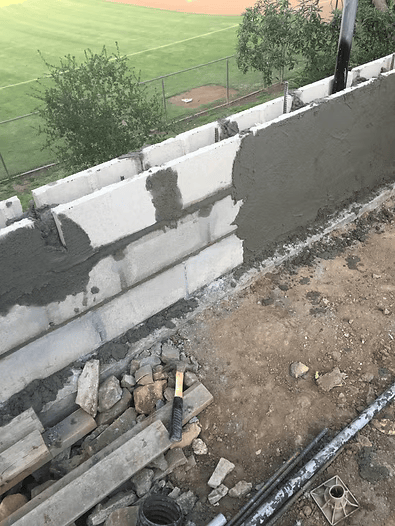 Construction of a concrete block wall, partially covered with wet cement. Beside a baseball field.
