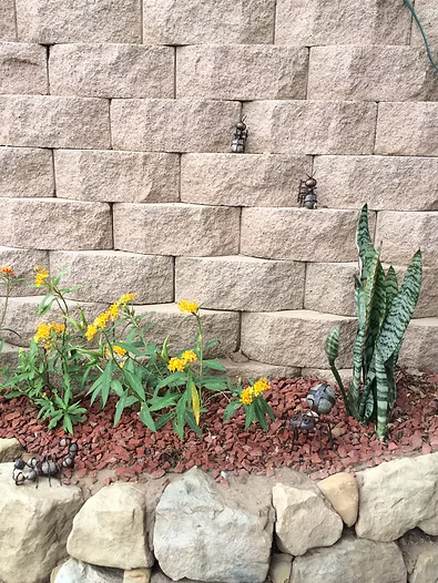 Beige block wall with flowers, plants, rocks in front. Two small statues sit on the wall.