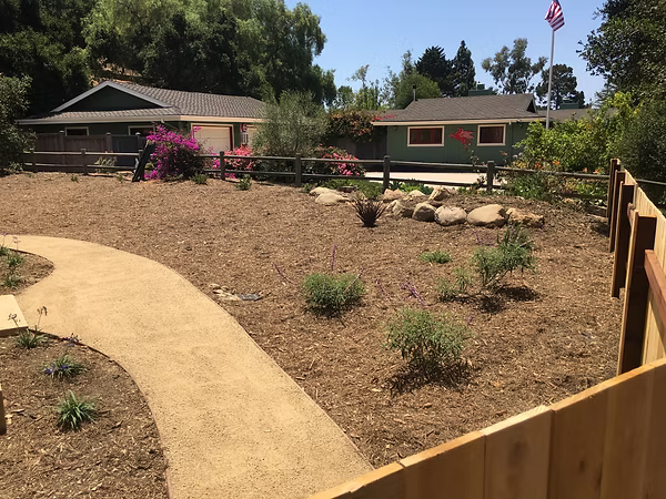 A tan path winds through a mulched garden toward two houses with pink flowers.