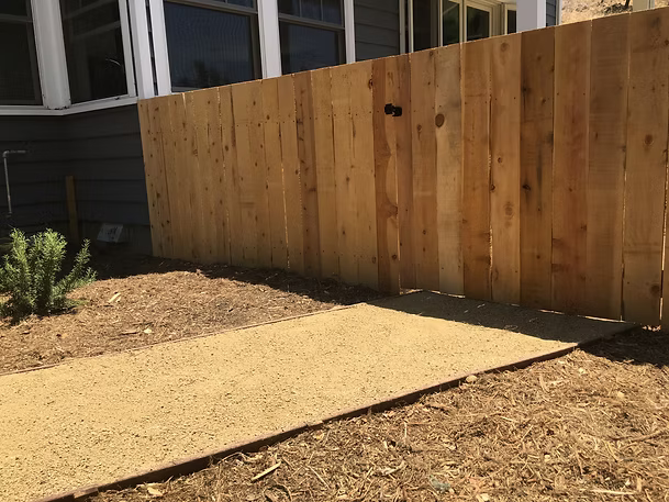 A wooden fence borders a gravel path next to a building with a blue-gray exterior.