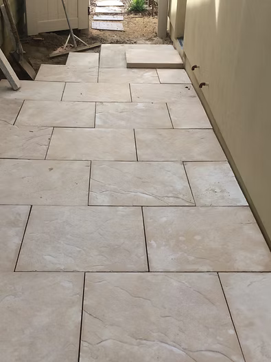 Beige rectangular patio tiles laid in a walkway next to a light beige wall.