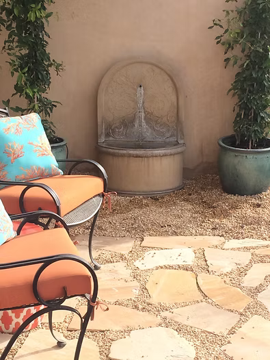 Outdoor patio with a fountain, chairs with orange cushions, plants, and stone pathway.