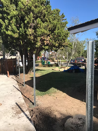 Metal fence posts installed alongside a concrete walkway, with a grassy yard and trees in the background.