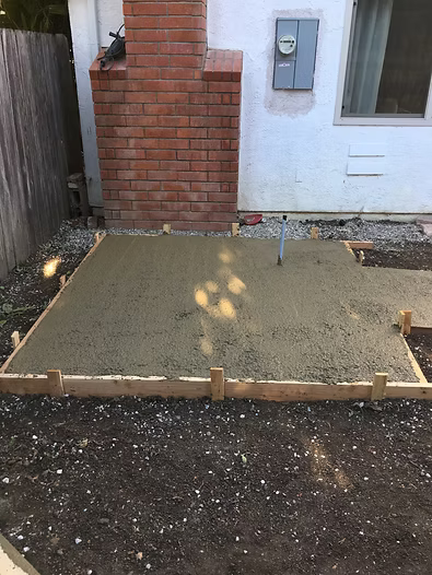 Freshly poured concrete patio with wooden formwork, near a brick chimney and electrical box.