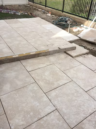 Pavers being laid on a patio; light beige squares with a level and construction materials visible.
