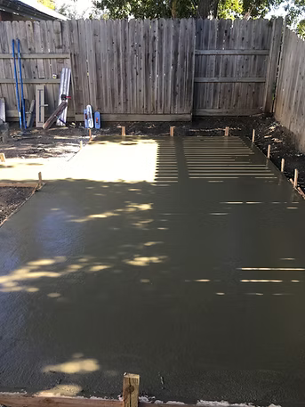 Freshly poured concrete slab in a fenced backyard, awaiting finishing.