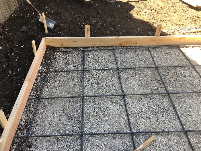 Concrete slab form with rebar grid, gravel base, and wooden frame, outdoors.