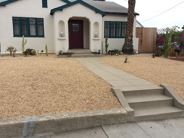 White house with brown door, gravel yard, concrete path and steps.