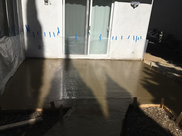 Freshly poured concrete patio in front of a white sliding glass door and wall. Blue tape marks are visible.