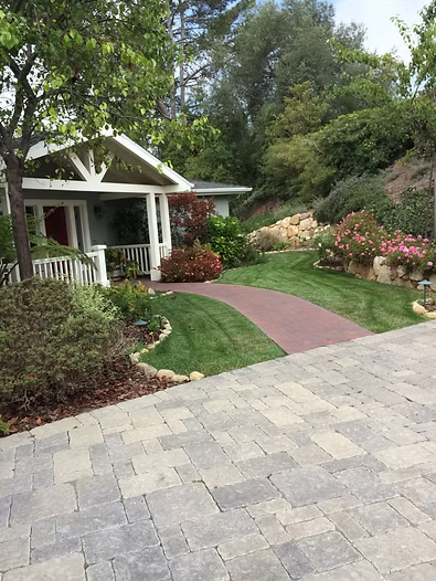 House with red brick pathway and green lawn, surrounded by landscaping and a paved driveway.