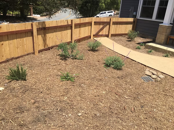 Wooden fence bordering a front yard with mulch, plants, and a concrete path.