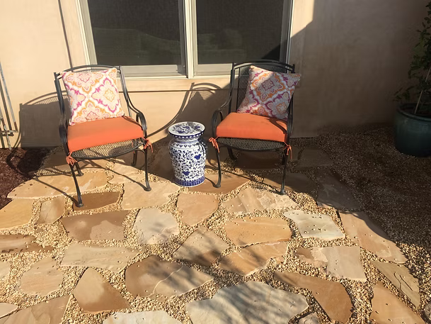 Two wrought iron chairs with orange cushions, decorative pillows, and a blue and white vase on a stone patio.