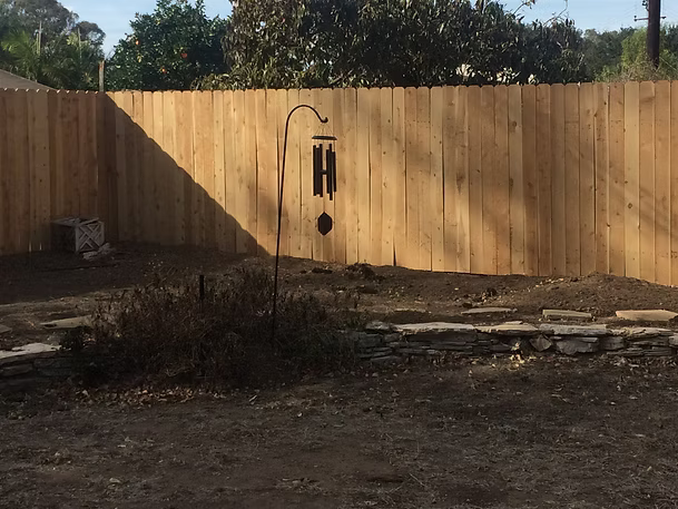 Wooden fence in a backyard with a wind chime and dried plants in front.