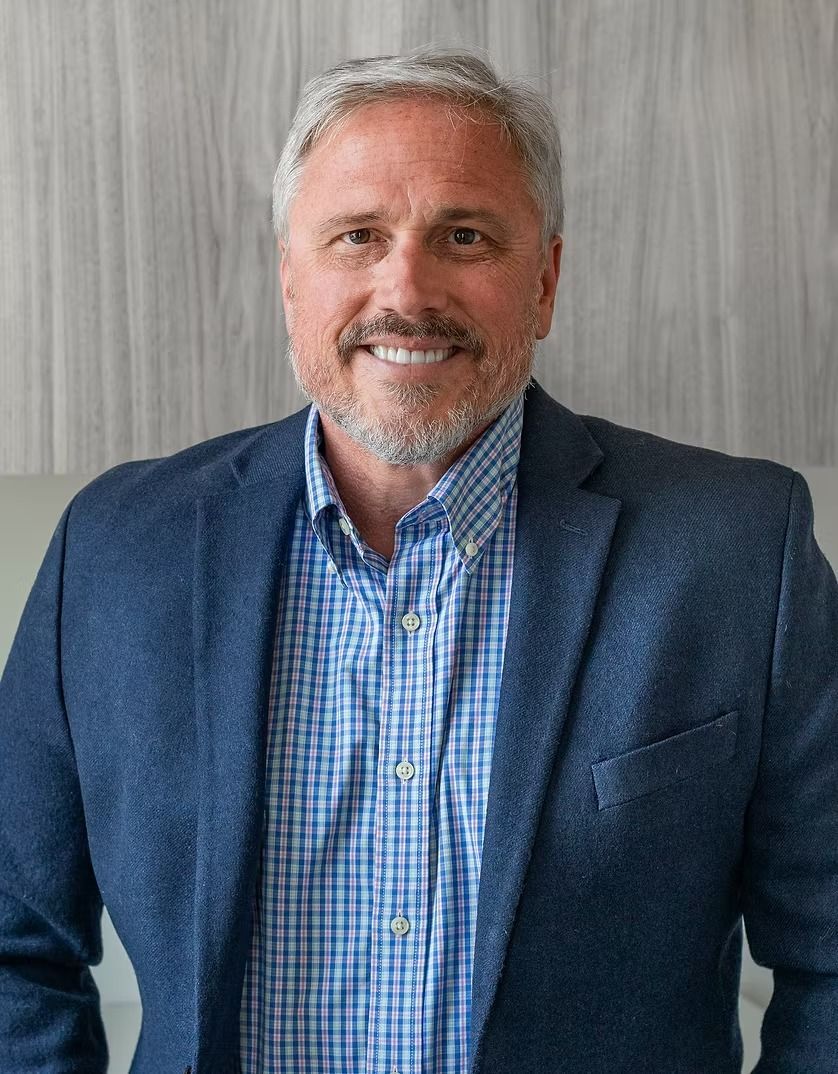 Man in a blue blazer and button-down shirt smiles, standing in front of a wood-paneled wall.