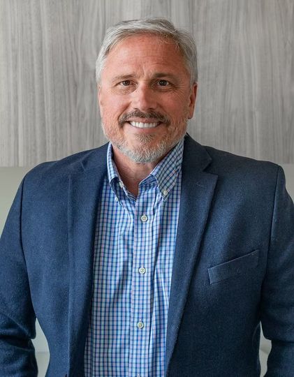 Man in a blue blazer and button-down shirt smiles, standing in front of a wood-paneled wall.