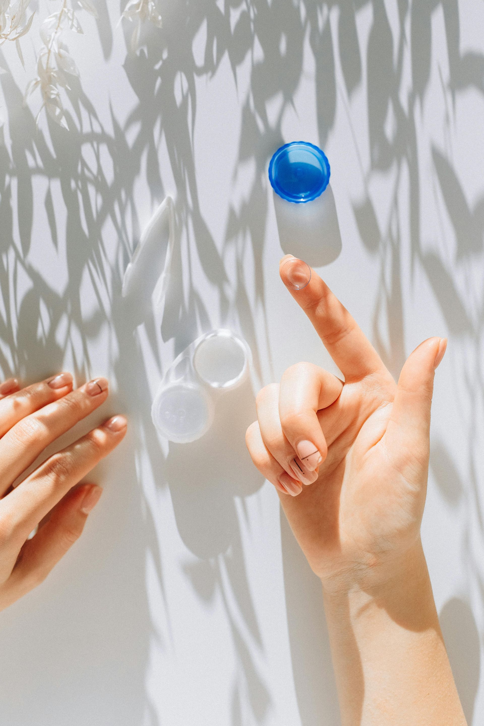 Hand balancing a blue contact lens case lid; white surface with shadows and two open cases.
