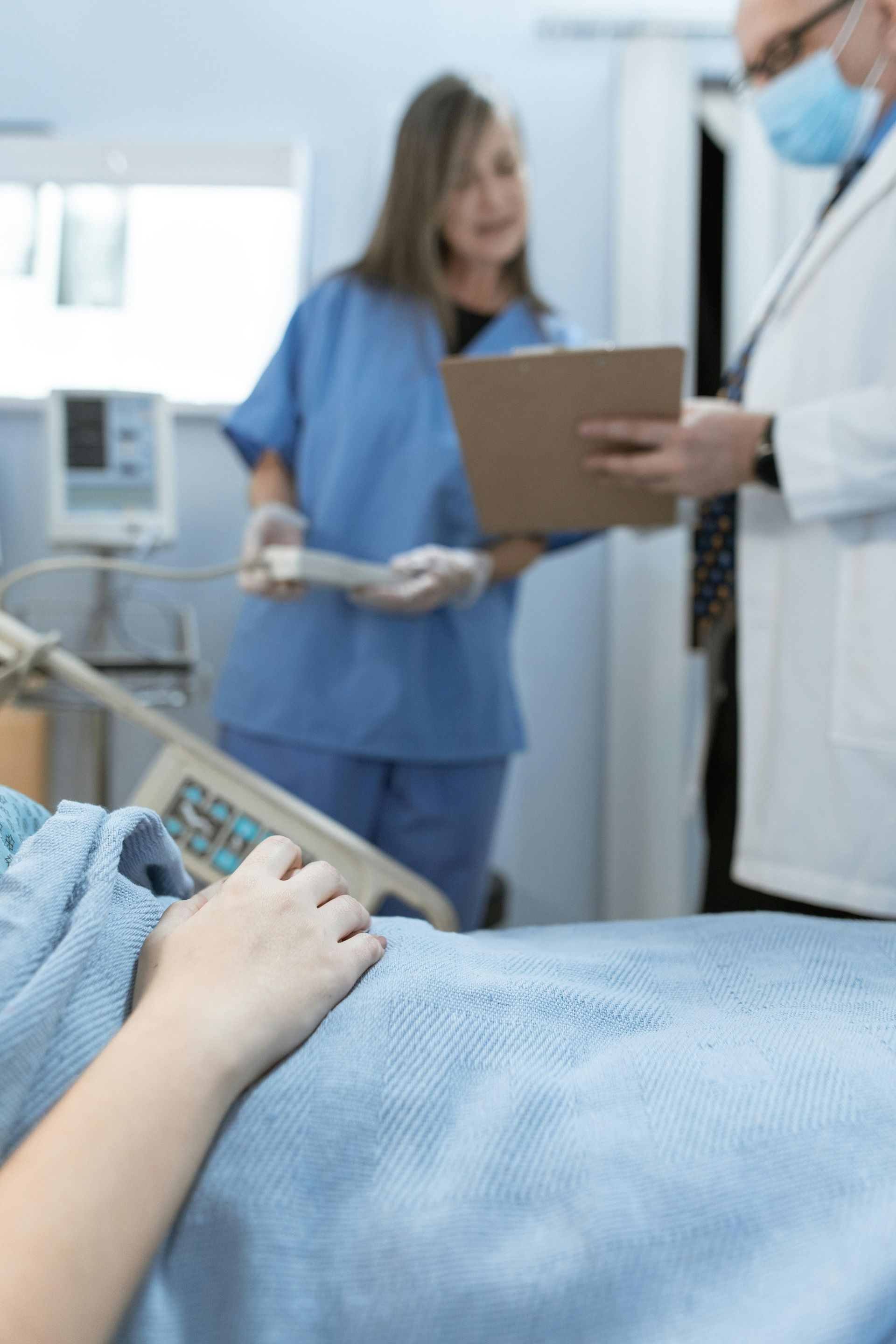 Patient's hand on bed, two medical professionals in scrubs and lab coat reviewing a clipboard in a hospital room.