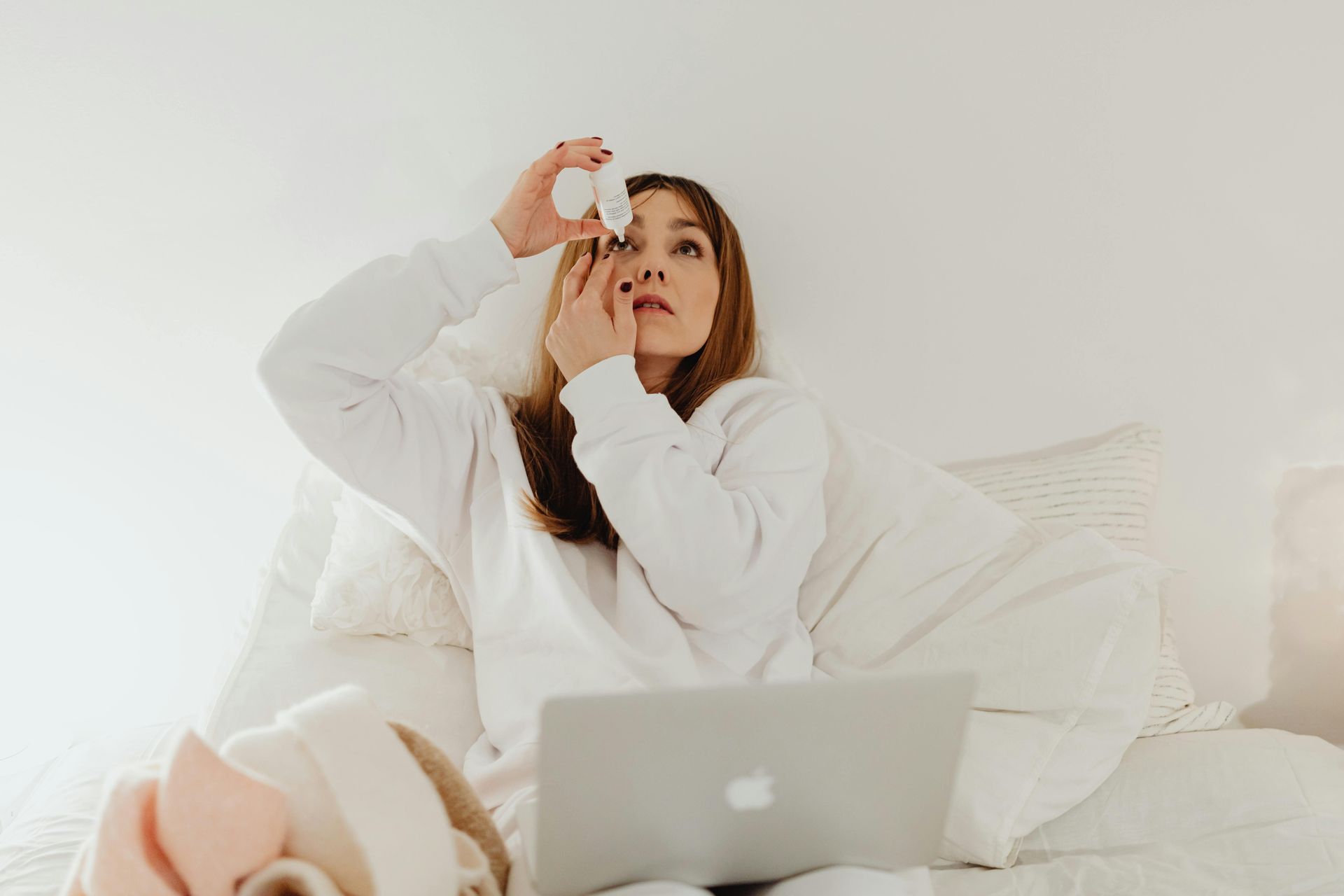 Woman in white shirt putting eye drops in, sitting on a bed with a laptop.