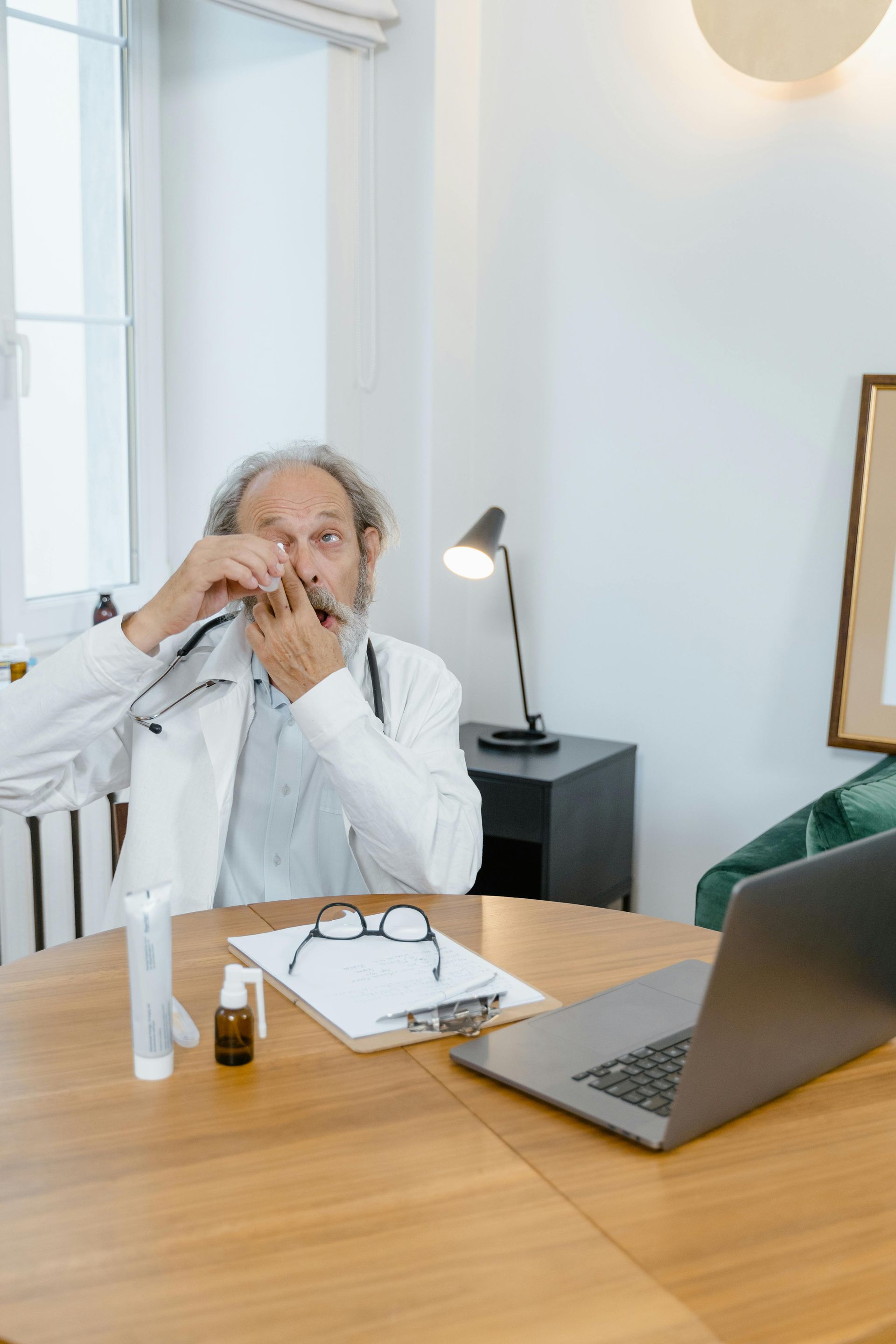 Doctor in white coat examining an inhaler, seated at desk with laptop, glasses, and medication.