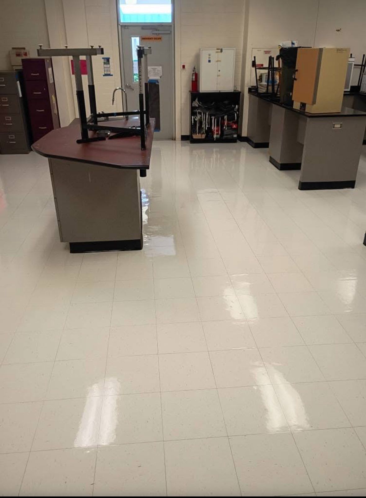 Empty science classroom with lab tables, sink, and emergency equipment. White tile floor reflects light.