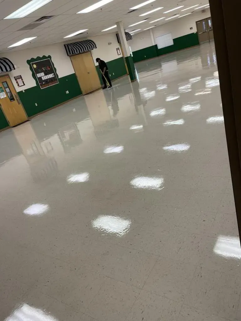Person sweeping a large room with a shiny floor. Green and white walls, white ceiling lights.