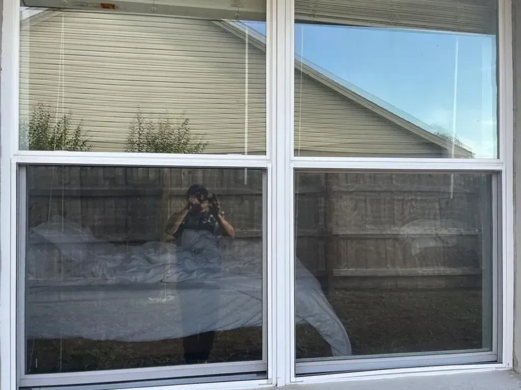 A person taking a photo reflected in a window. The window overlooks a wooden fence and cloudy sky.