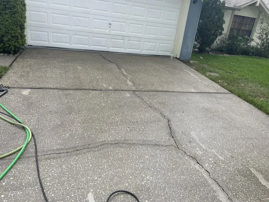 Cracked concrete driveway with a white garage door in the background. A green hose is visible on the left.
