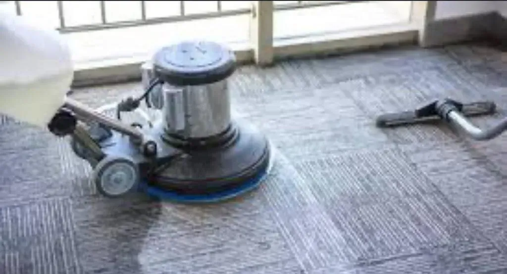A machine scrubbing and cleaning a carpeted floor near a window.