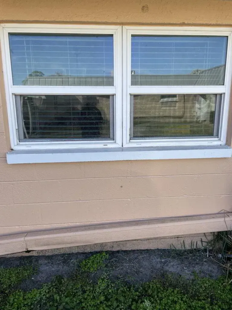 Two white-framed windows with closed blinds on a peach-colored wall, below a slanting concrete base and green grass.