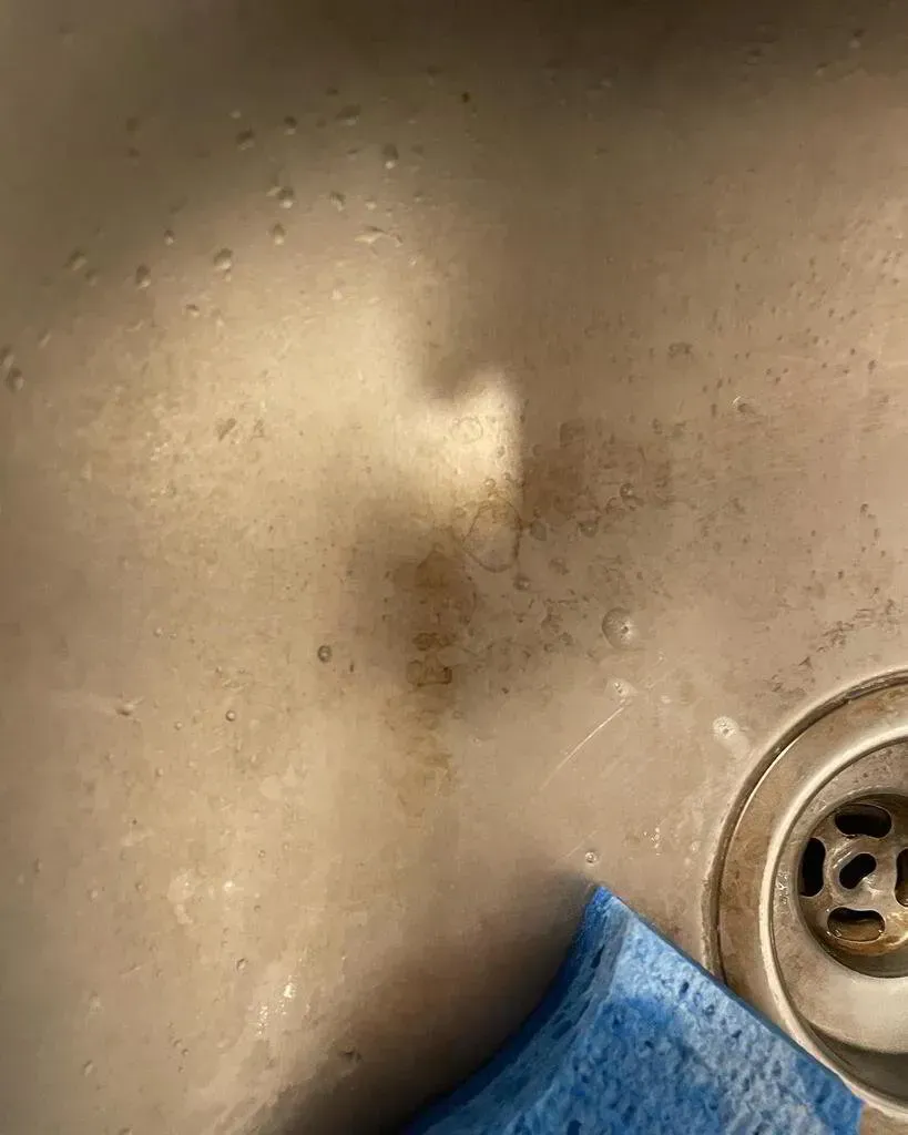 Stainless steel sink with brown stains, a blue sponge, and a drain.