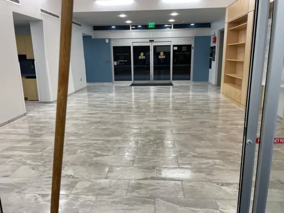 Empty, tiled lobby with automatic doors, blue wall, and partially built shelving. A broom leans in the foreground.