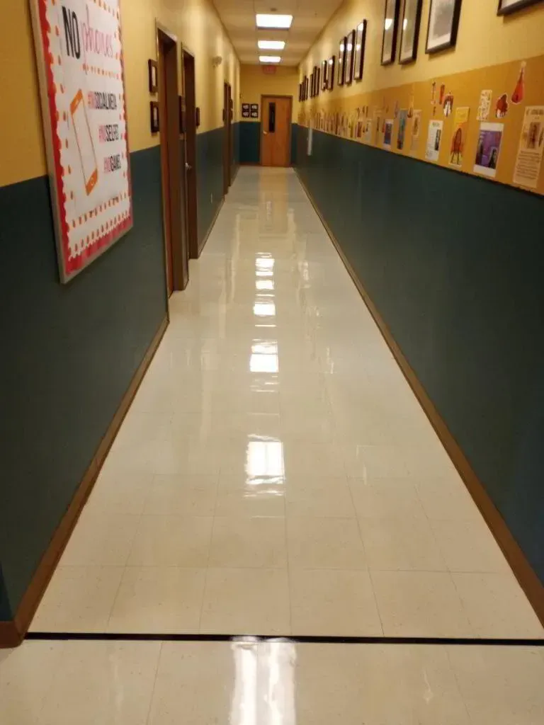 Long, empty school hallway; shiny white floor, dark green and gold walls, doors on the left, bulletin board on the right.