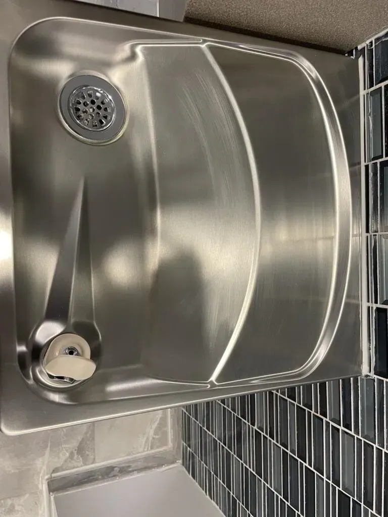 Stainless steel sink with drain and curved basin, set into countertop with dark tile backsplash.