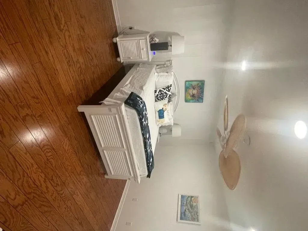 Bedroom with white bed, wooden floors, nightstand, ceiling fan, and art on the walls.