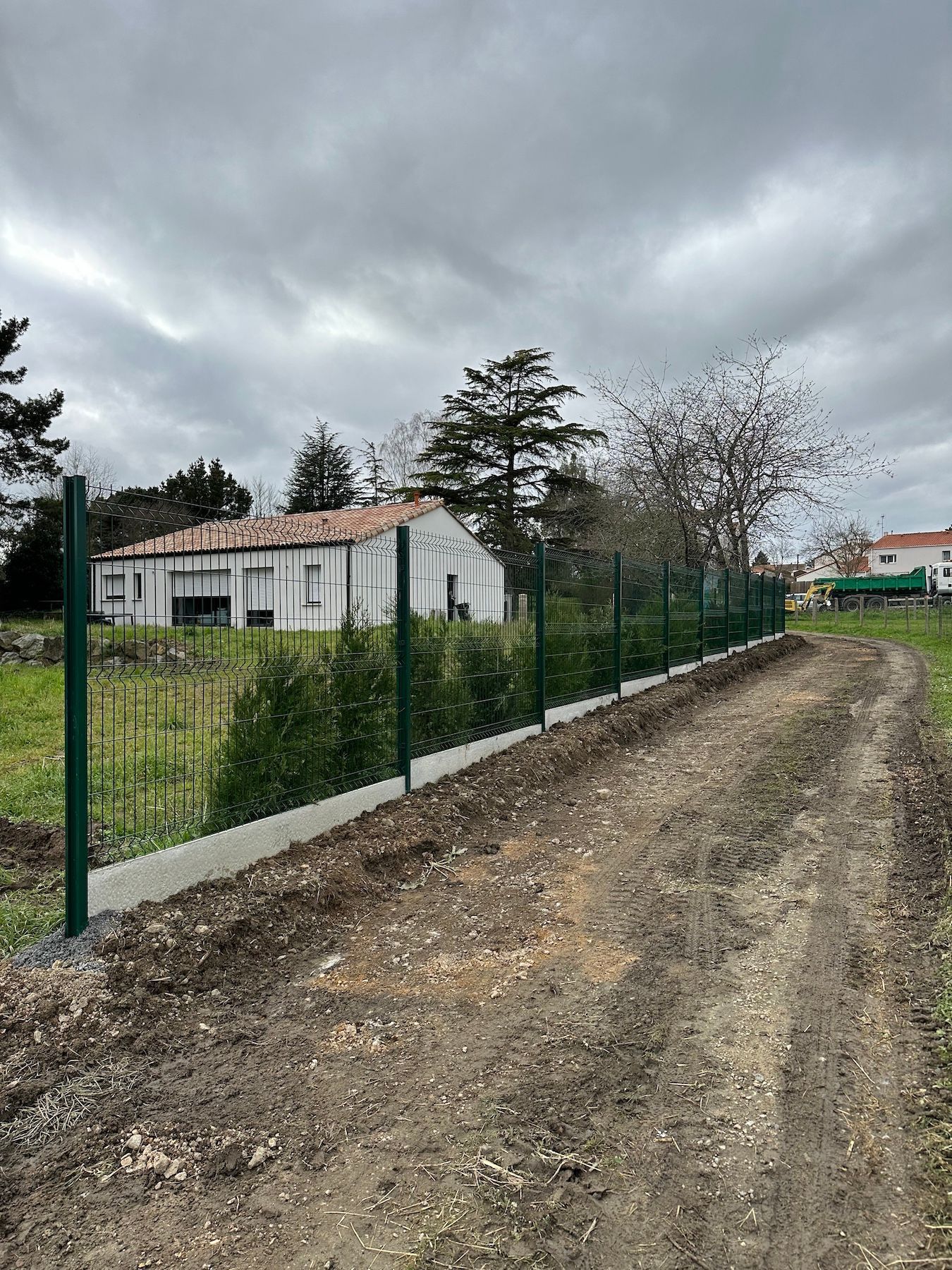 Une clôture verte entoure un chemin de terre devant une maison.