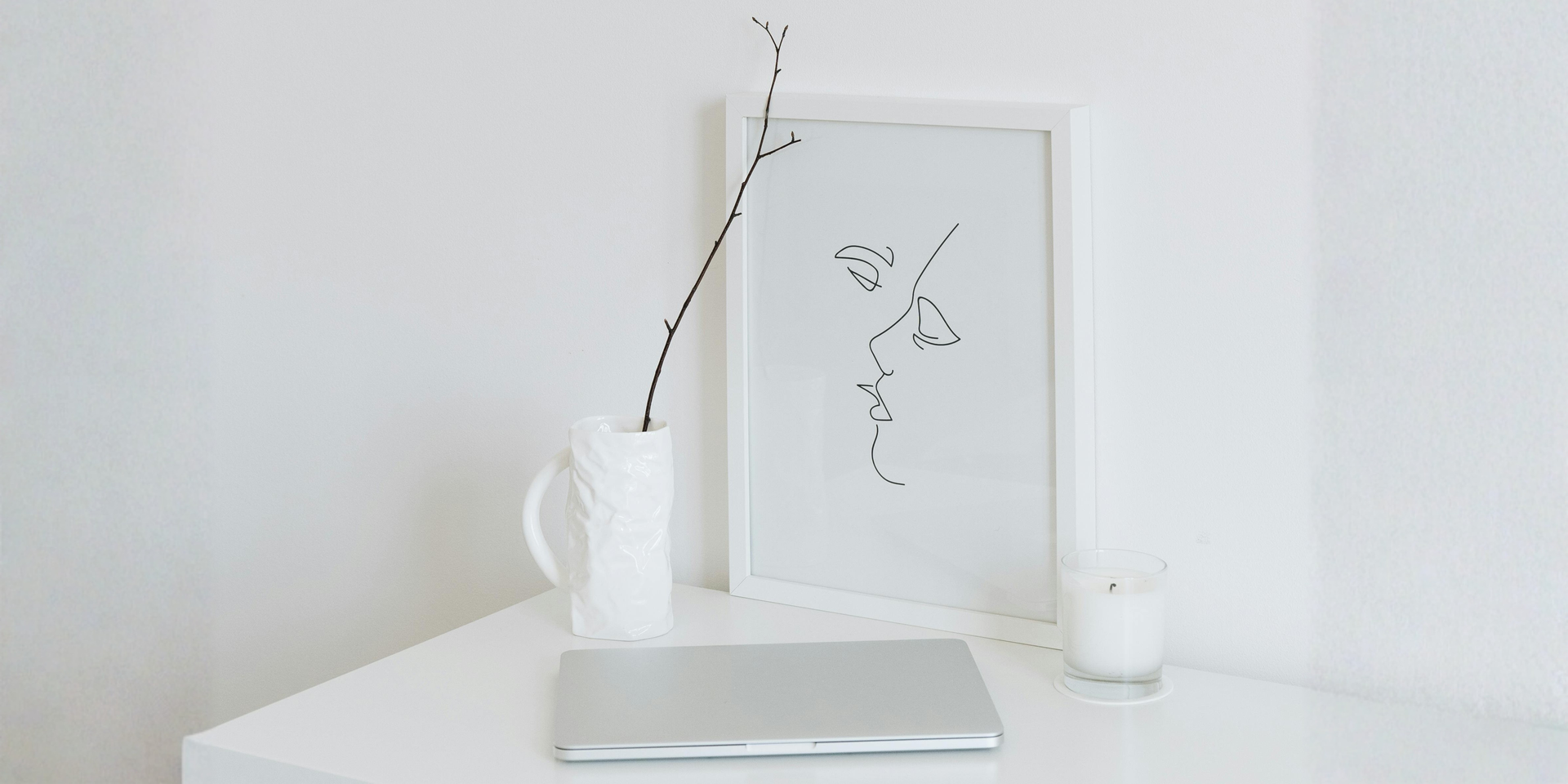 White desk with a laptop, vase with a branch, art, and a candle, all in a minimalist white room.