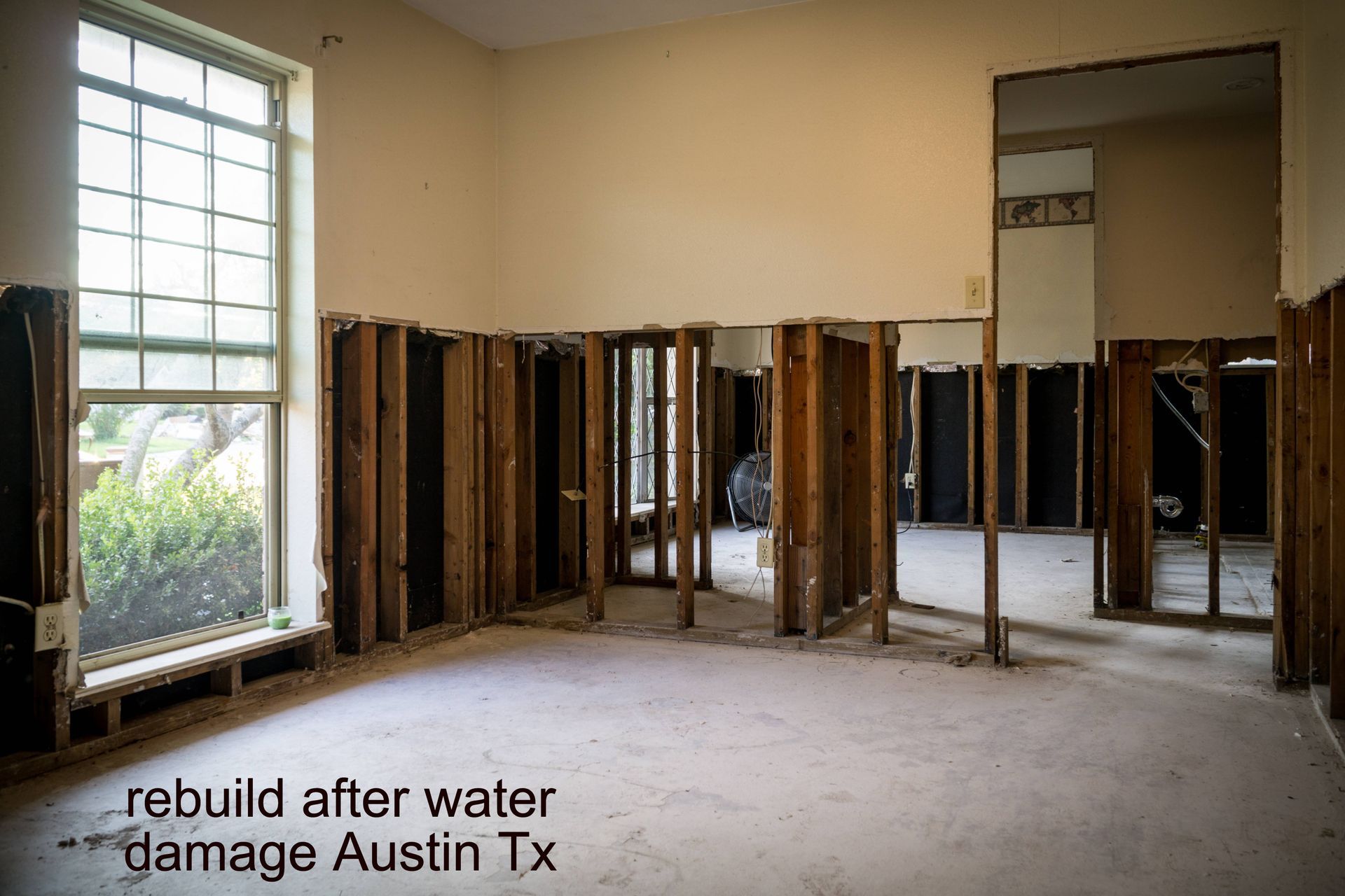 Interior room with water damage, exposed framing, walls removed. Large window, bare concrete floor.