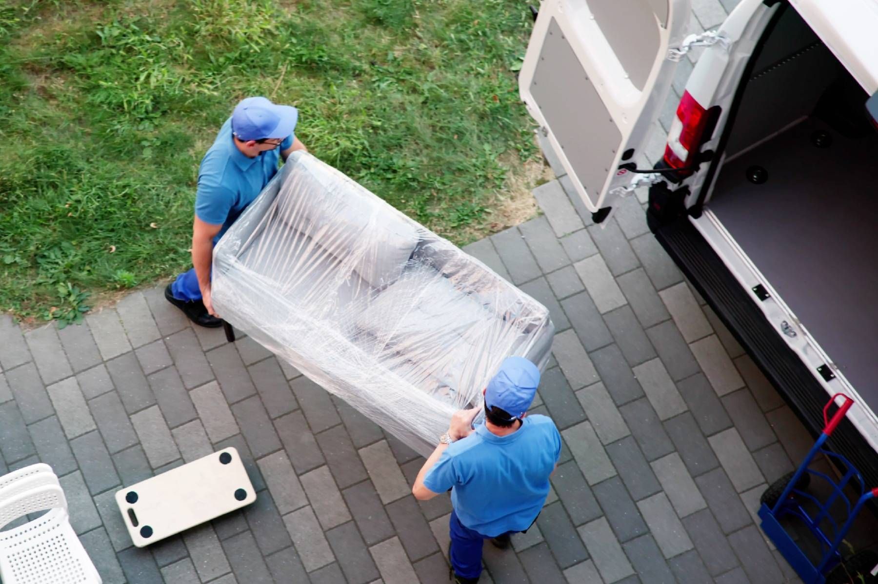 Two movers in blue shirts load a wrapped sofa into a white van on a brick patio.