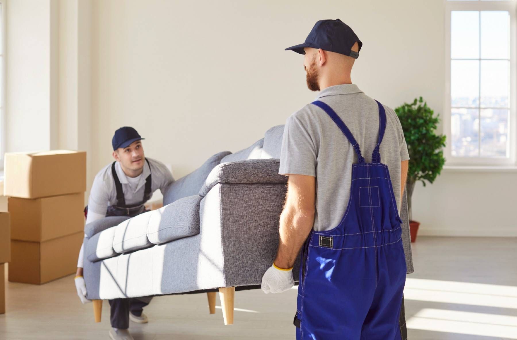 Two movers in overalls carrying a grey couch inside a room with boxes and a window.